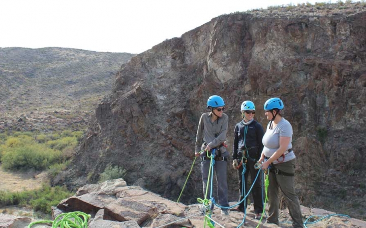 a group of three outward bound students wearing rock climbing gear prepare to rappel down into a canyon